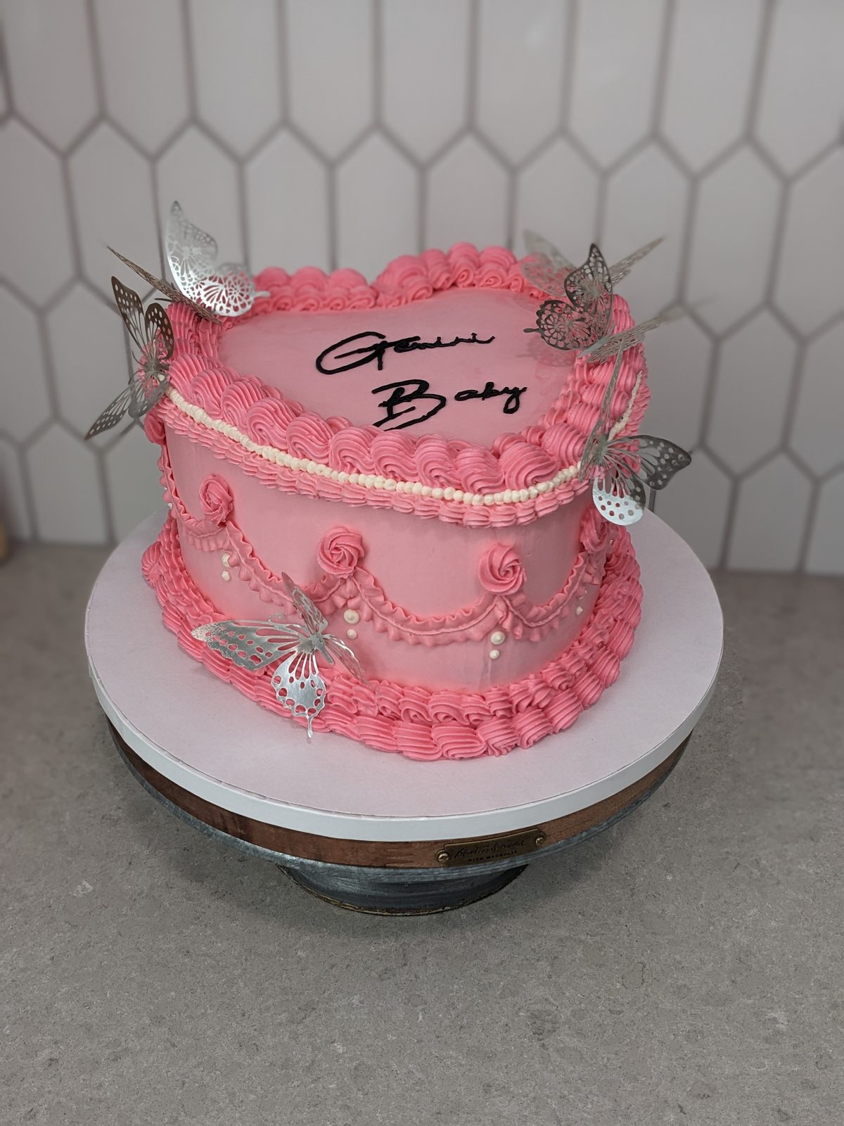 Pink heart-shaped buttercream cake piped with swags and roses, topped with silver butterflies and Gemini Baby in script