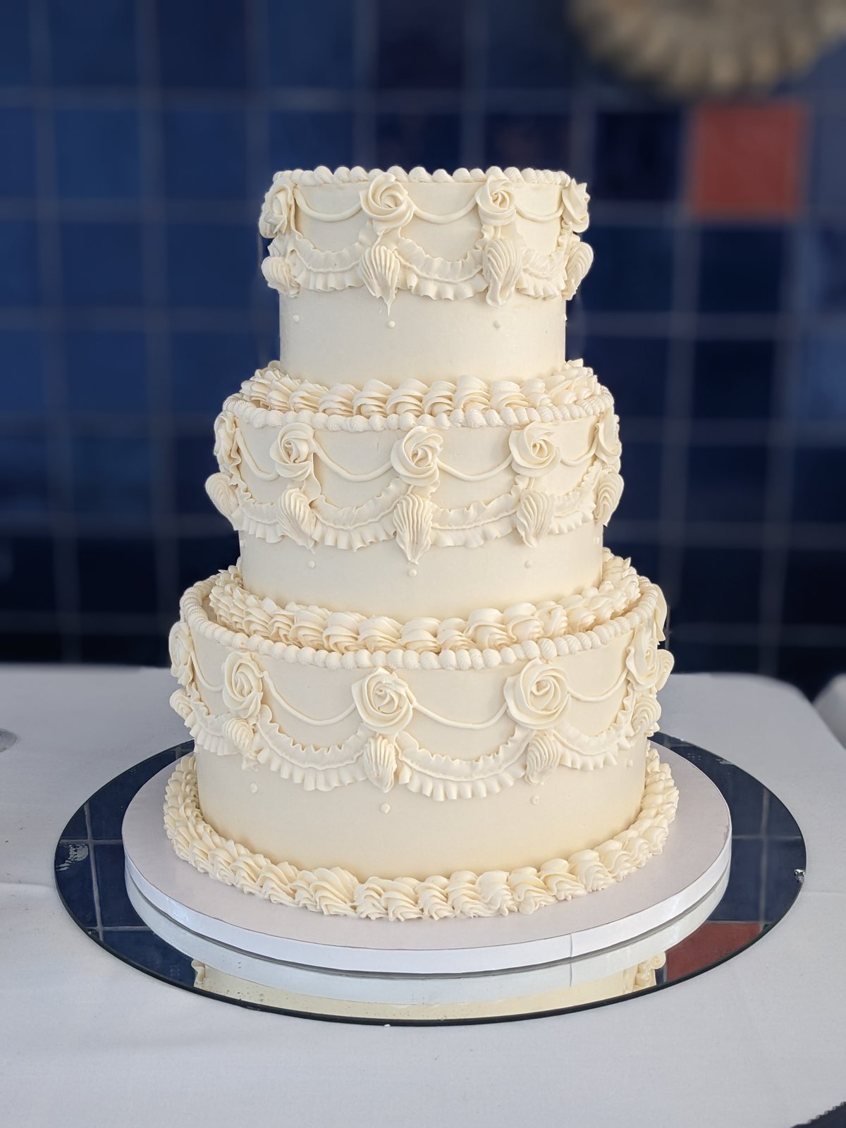 Three-tier ivory buttercream wedding cake piped with swags, roses and shells, against a deep blue tiled wall